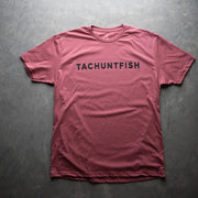 A dusty rose pink short-sleeve t-shirt with the word 'TACHUNTFISH' printed in black across the chest, laid flat on a dark gray textured surface.